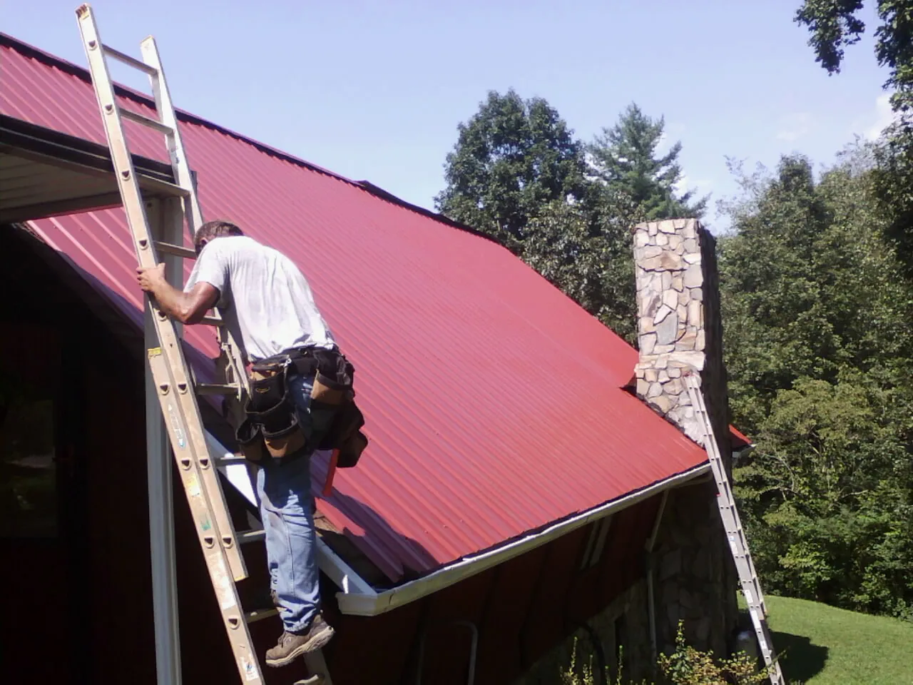 Roofer on a ladder beside a red metal roof and stone chimney — Soffit & Fascia Repair by Roofing Brothers in Malin